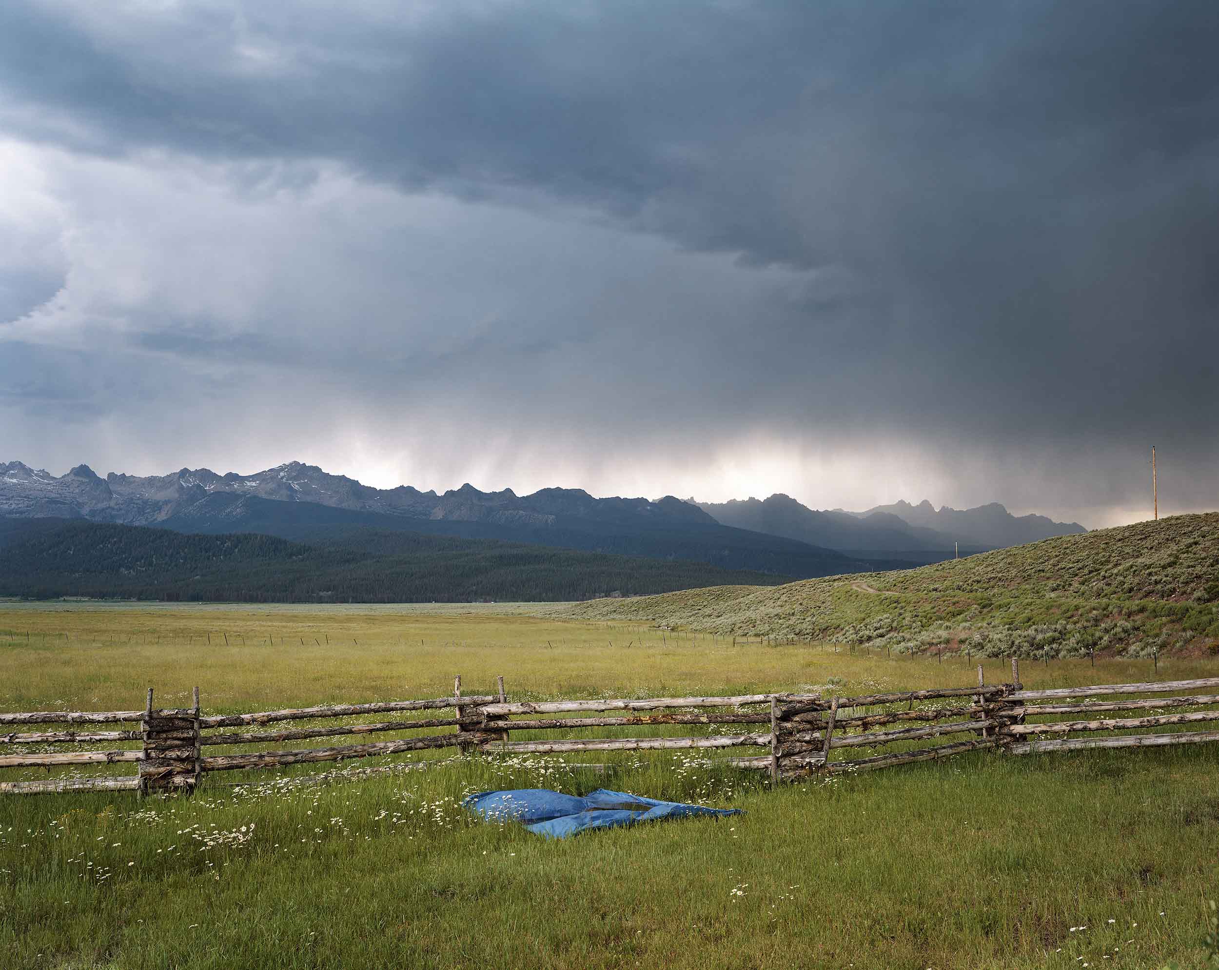 Green field with wooden fence in foreground and mountains in background, dark clouds dropping rain overhead