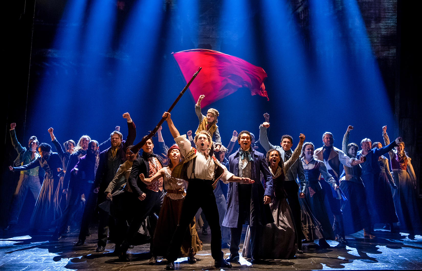 The "Les Miserables" ensemble appears on stage, with a man holding a gun in front and a red flag in the background.