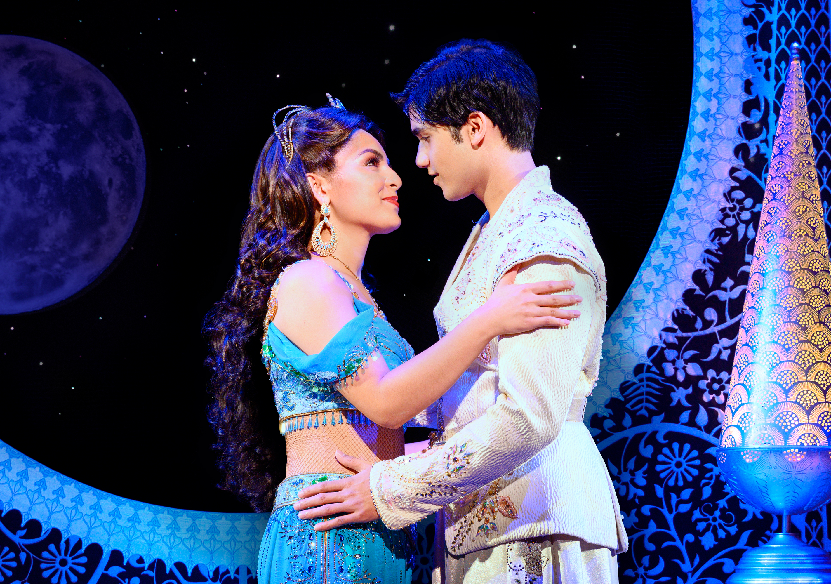 A young woman and young man hold each other and look into each other's eyes in a palace room in "Aladdin."