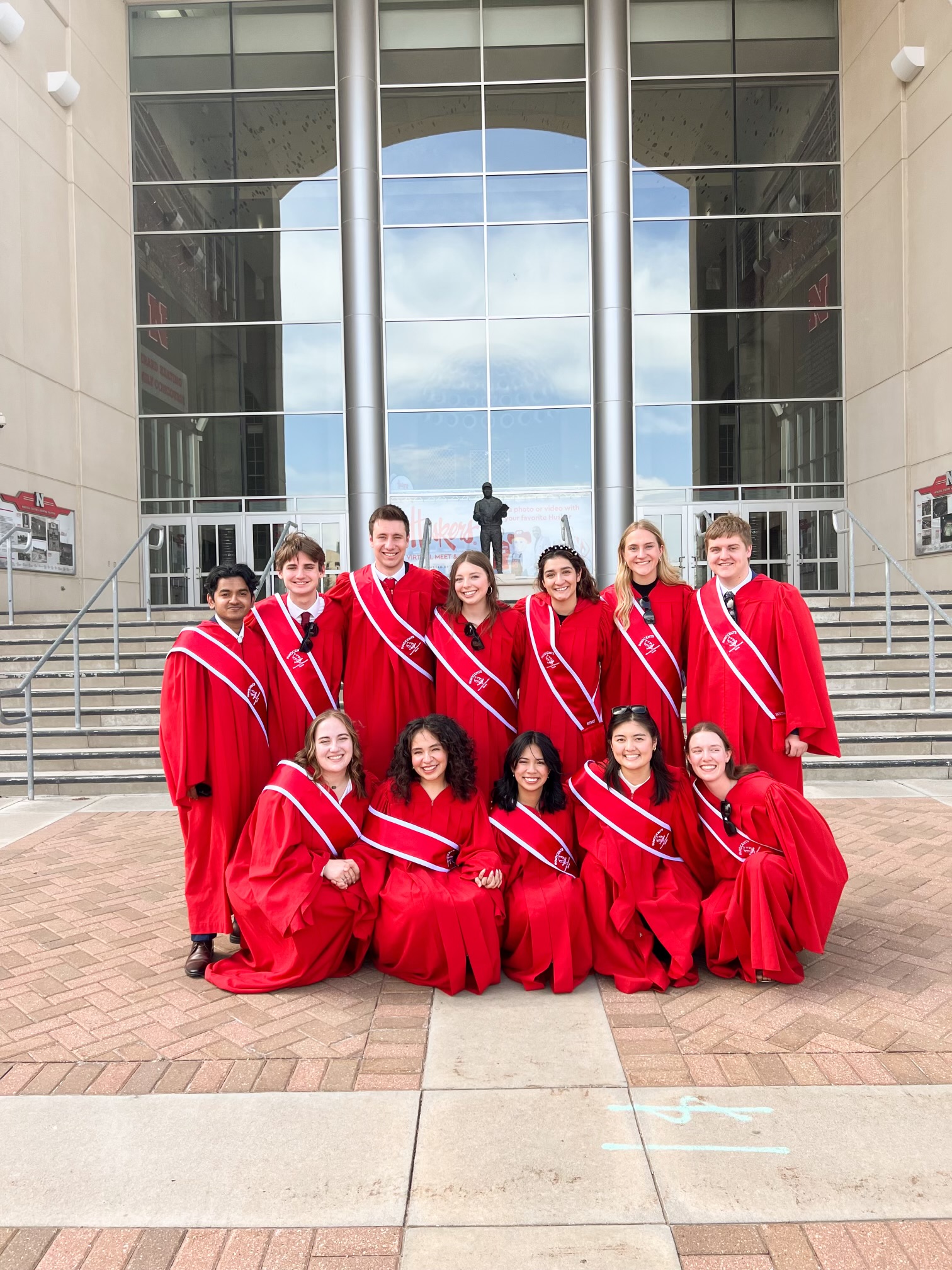 Twelve college students in red robes with red sashes pose on the plaza east of Memorial Stadium.