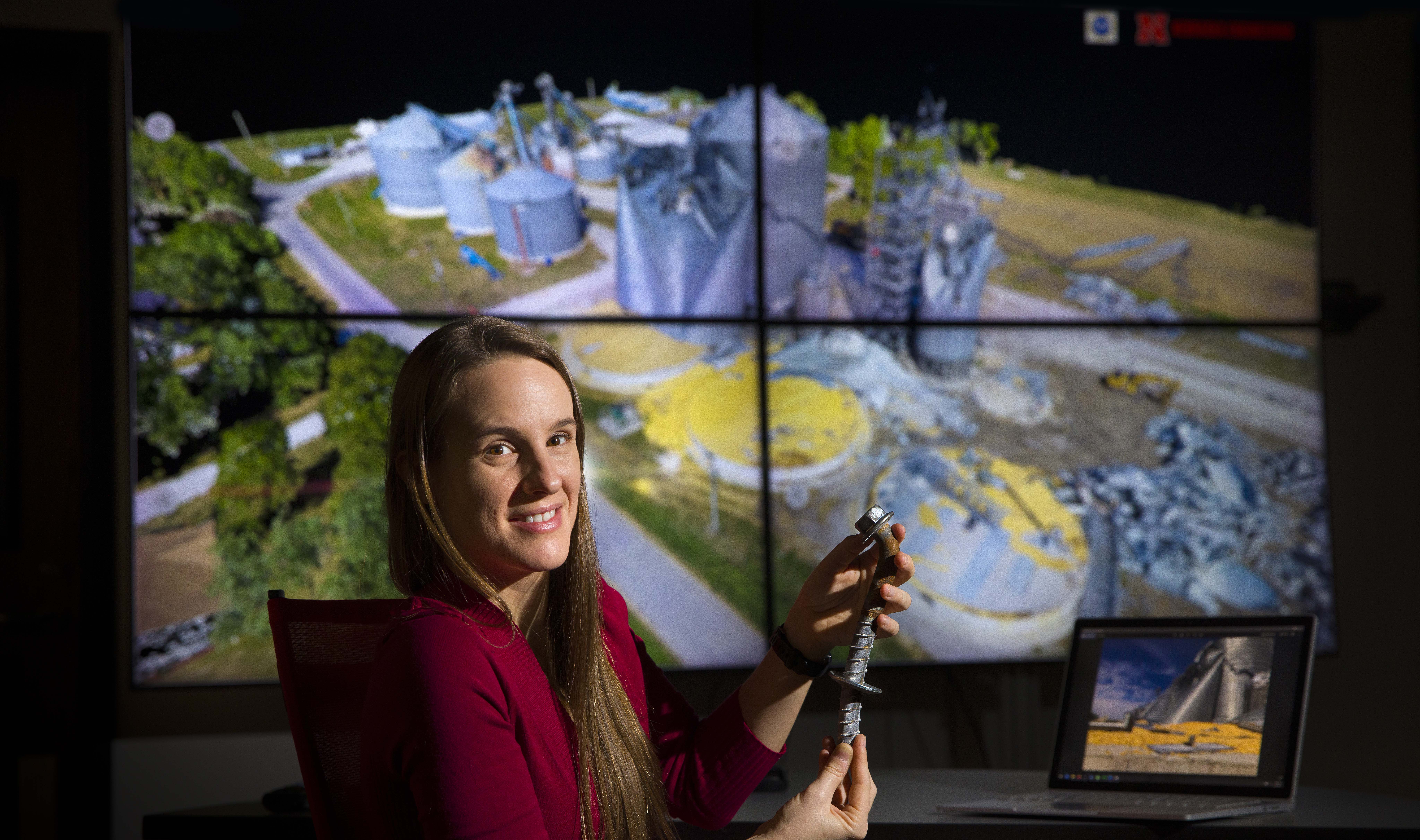 Christine Wittich holds a damaged anchor bolt in front of a photo of damaged steel grain bins.