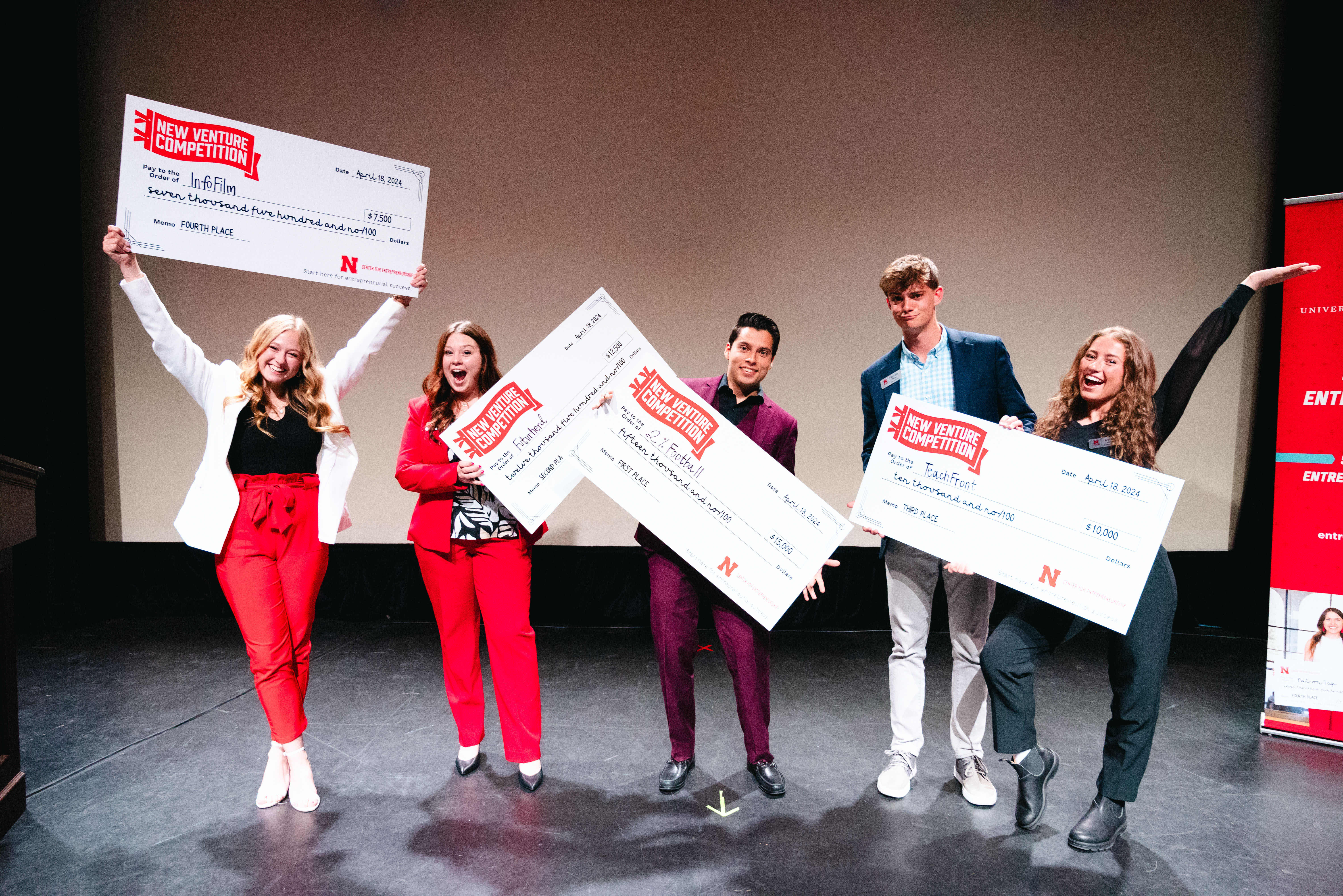 Three young women and two young men hold giant checks on a stage.