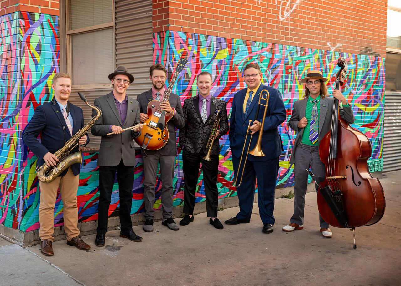 Six men with jazz instruments stand in front of a colorful mural on a building.