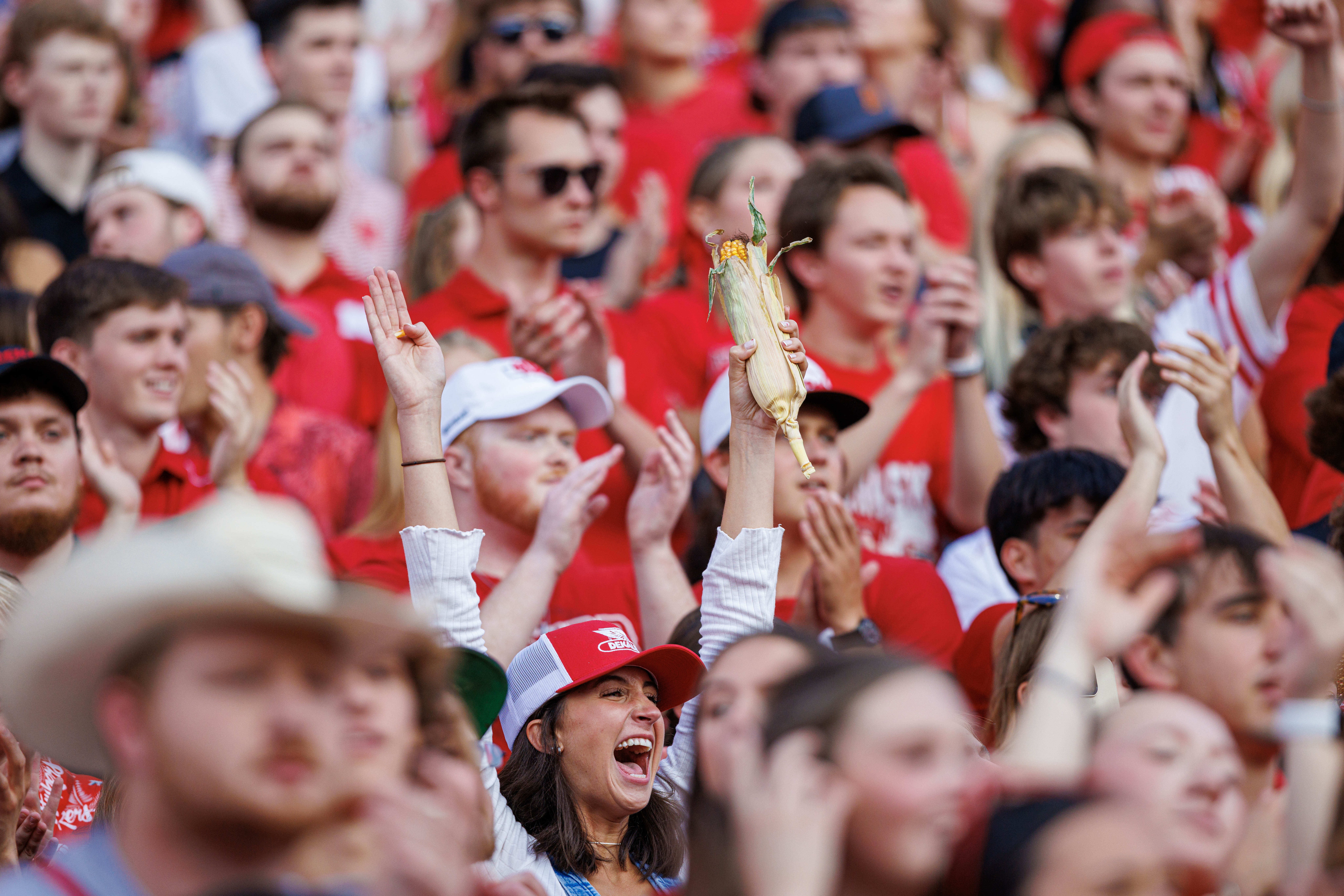 A woman holds her arms above her head, while holding an ear of corn, amid a crowd of Husker fans.