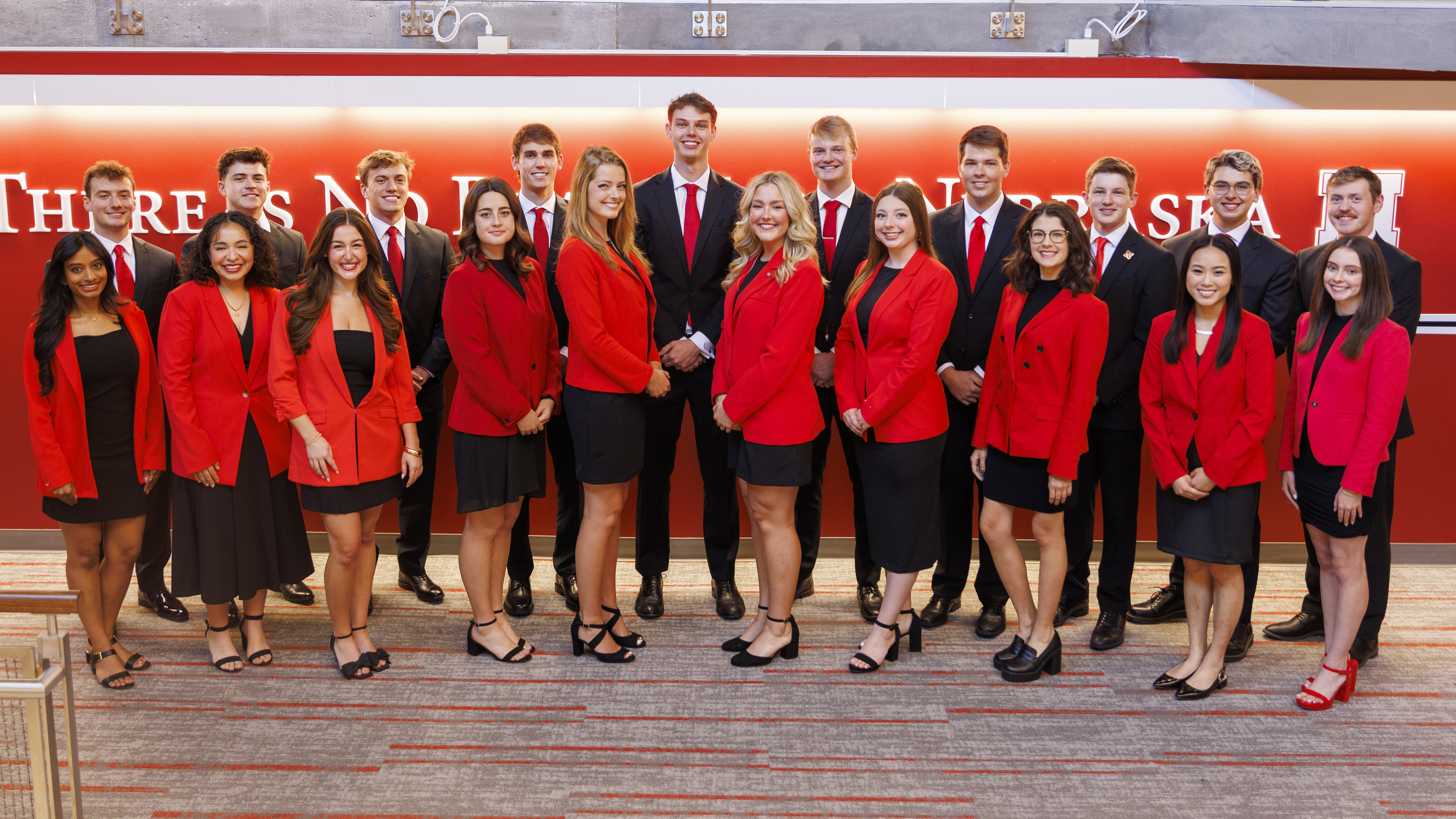 Twenty Husker seniors, wearing red and black outfits, pose in front of a wall that reads "There is no place like Nebraska."