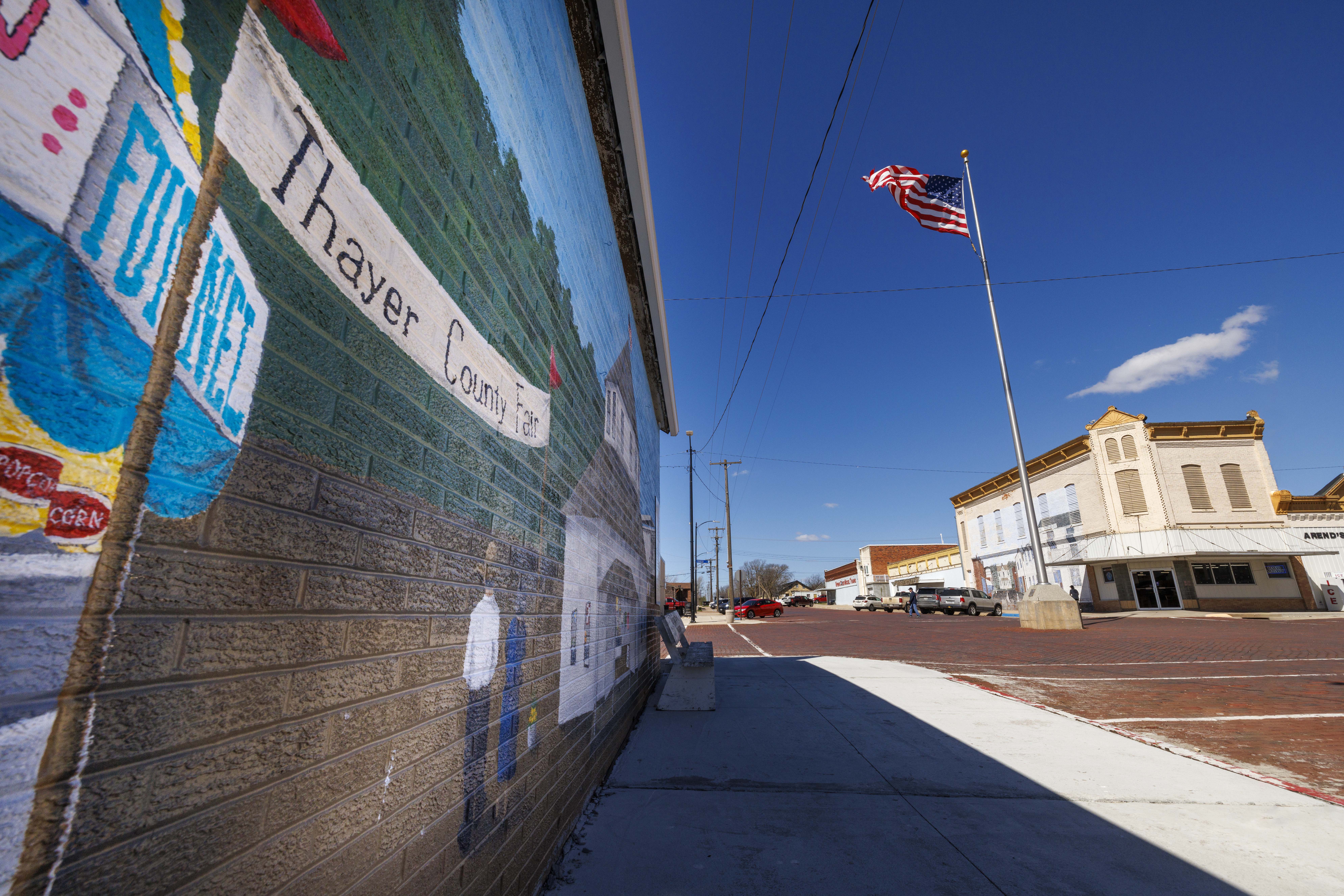 A Thayer County mural is painted on a brick wall in downtown Deshler, Nebraska. An American flag flies in the background.