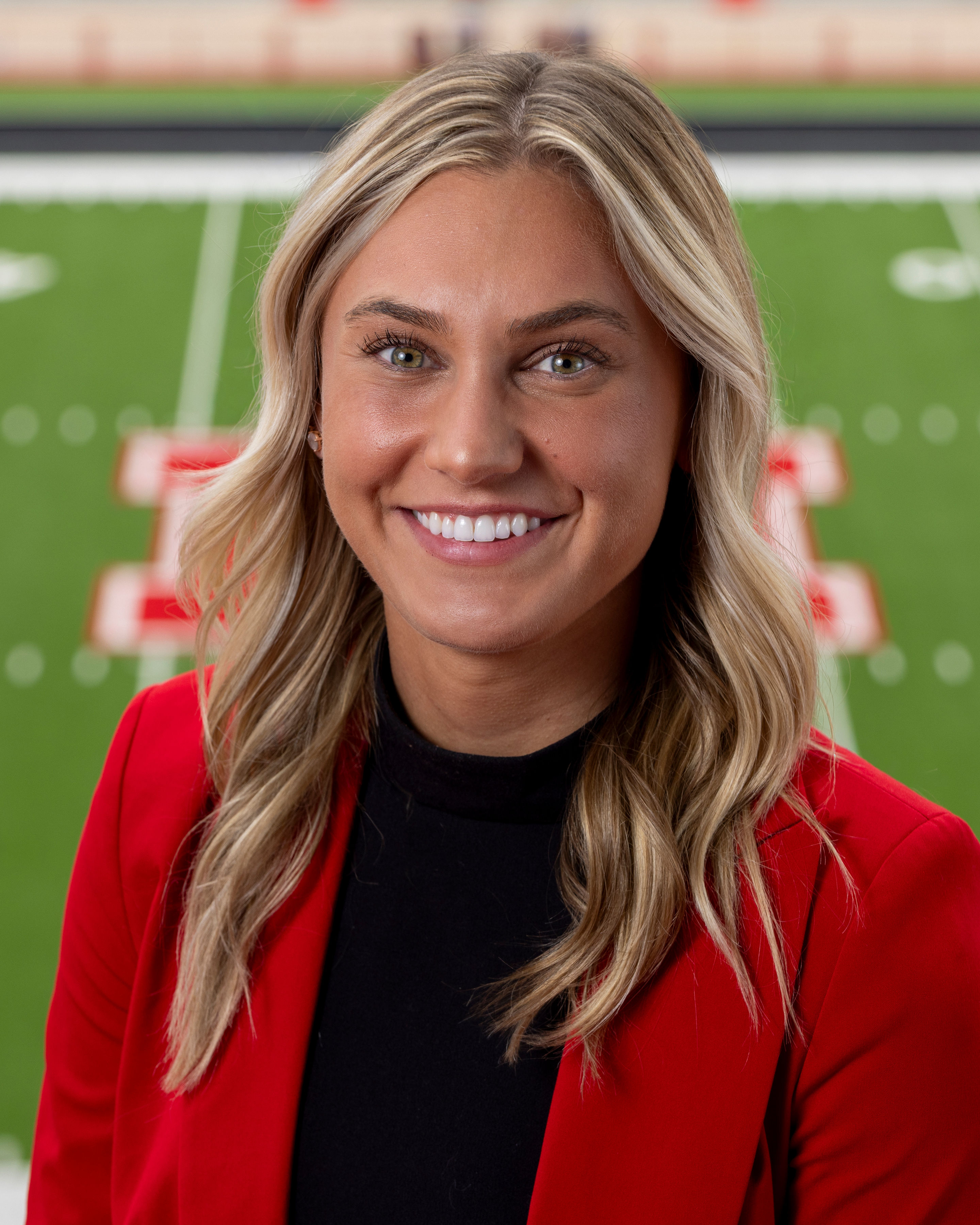 Color portrait of Ava Hollingsworth, wearing a red blazer over a black shirt, with Tom Osborne Field in the background.