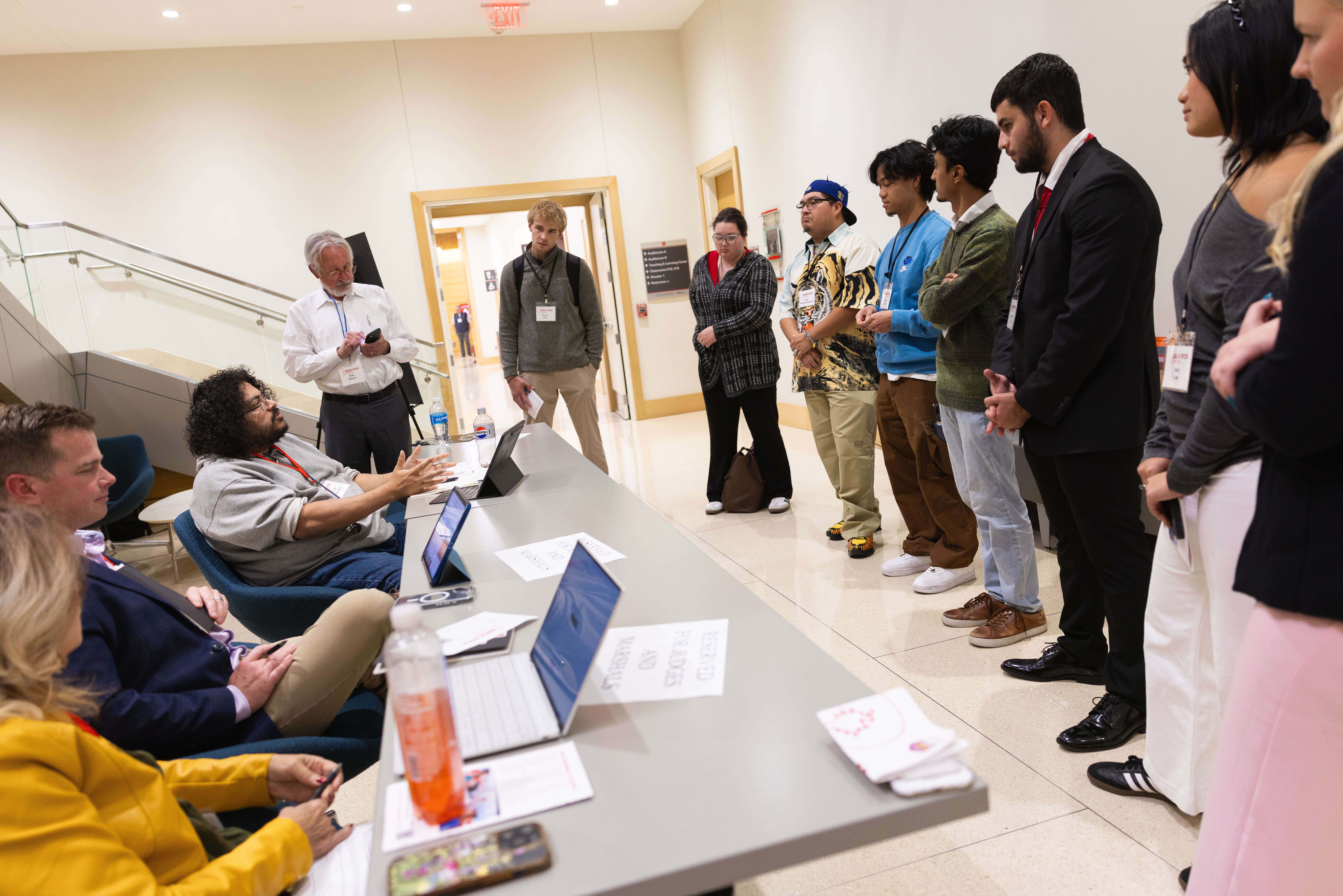 Eight students — participants in this year's 3-2-1 Quick Pitch competition — stand in front of a table of three judges in a hallway in Howard L. Hawks Hall.