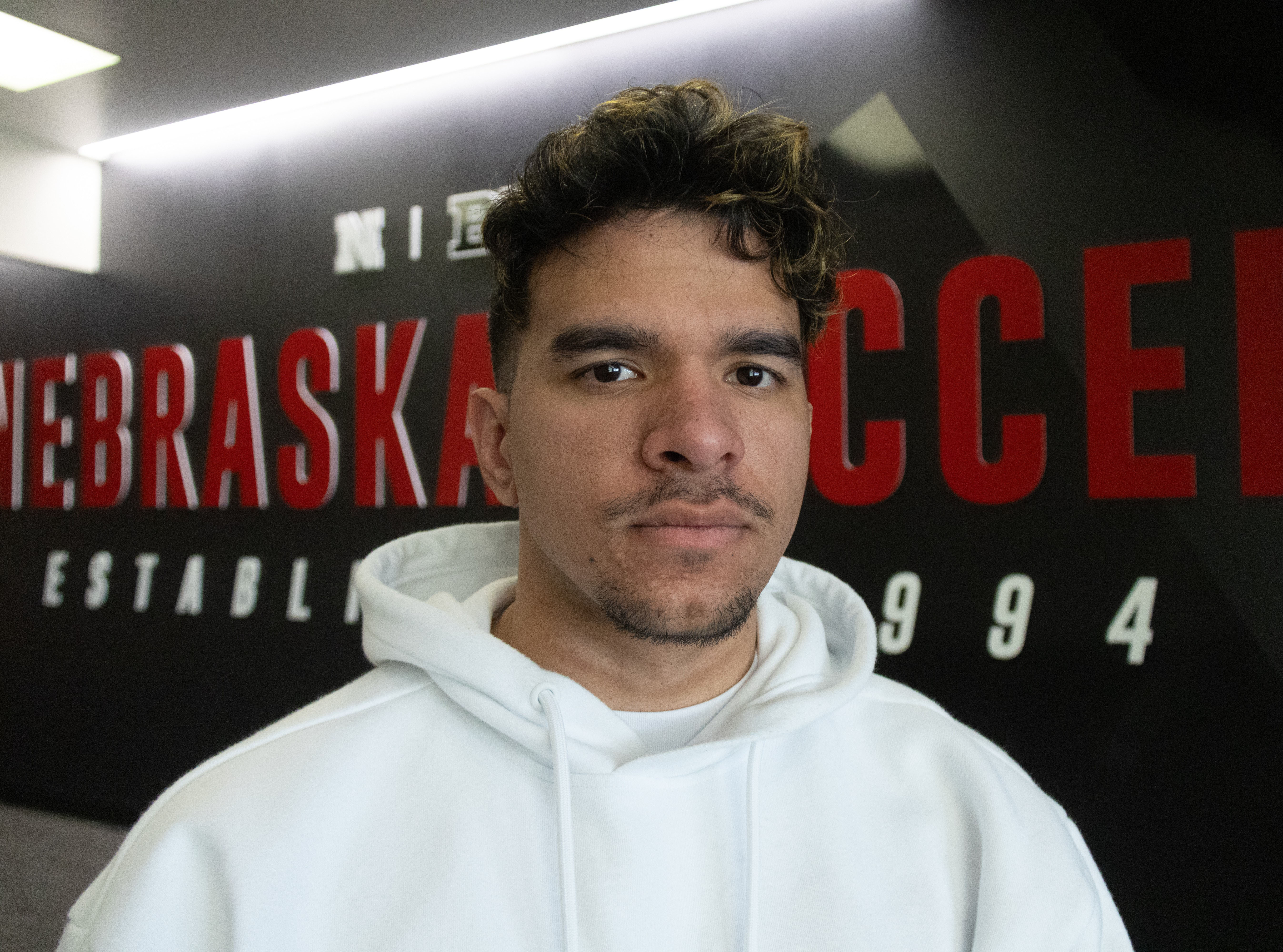 Color portrait of Kevin Artiles Fernandez, senior accounting and sports media and communication major at the University of Nebraska–Lincoln. He is wearing a white hoodie, standing in front of a wall that reads "Nebraska Soccer, Established 1994."