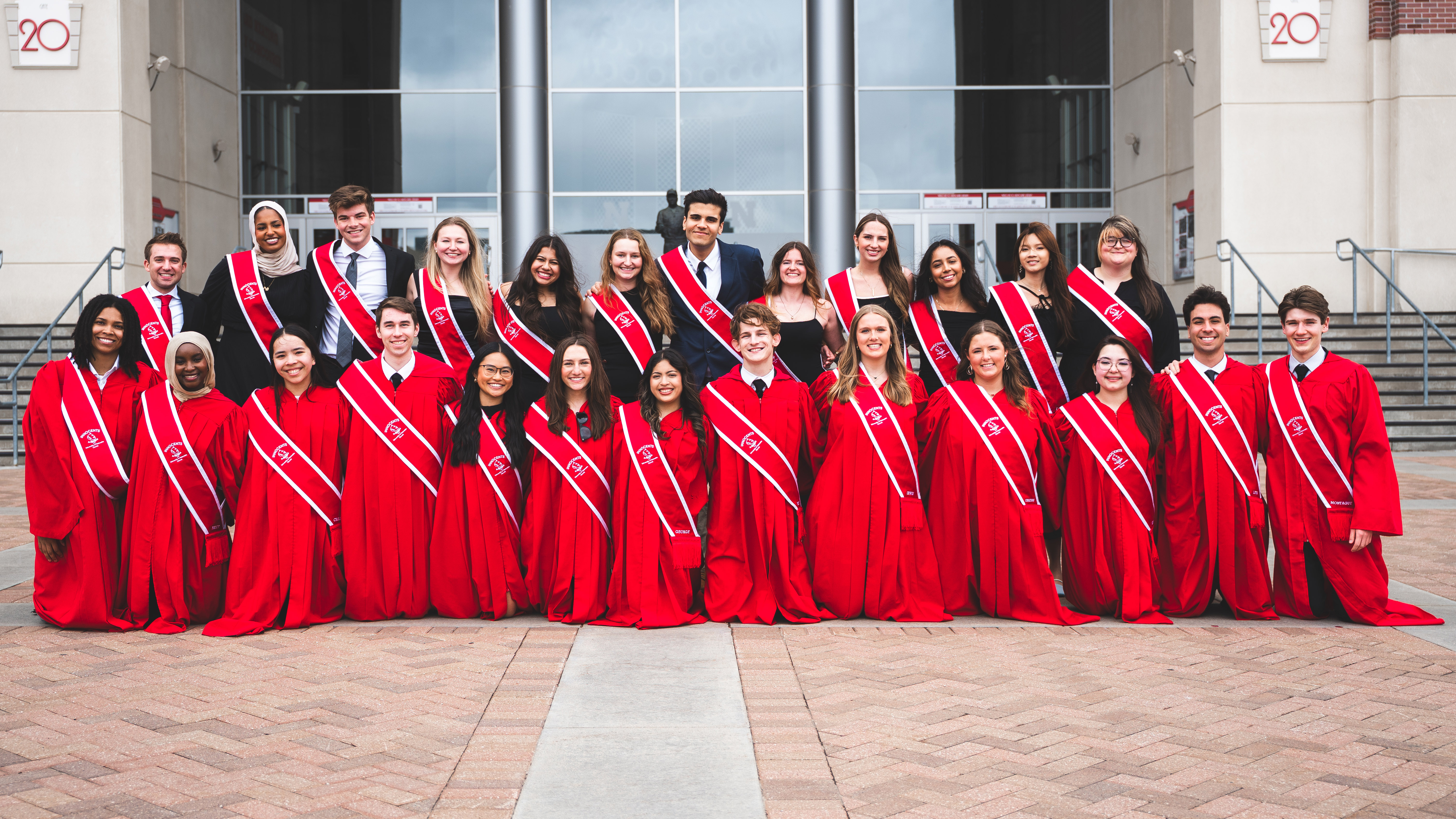Twenty-five University of Nebraska–Lincoln students — 12 in formal attire and 13 in red robes, all wearing red sashes — pose for a group photo on the east side of Memorial Stadium.