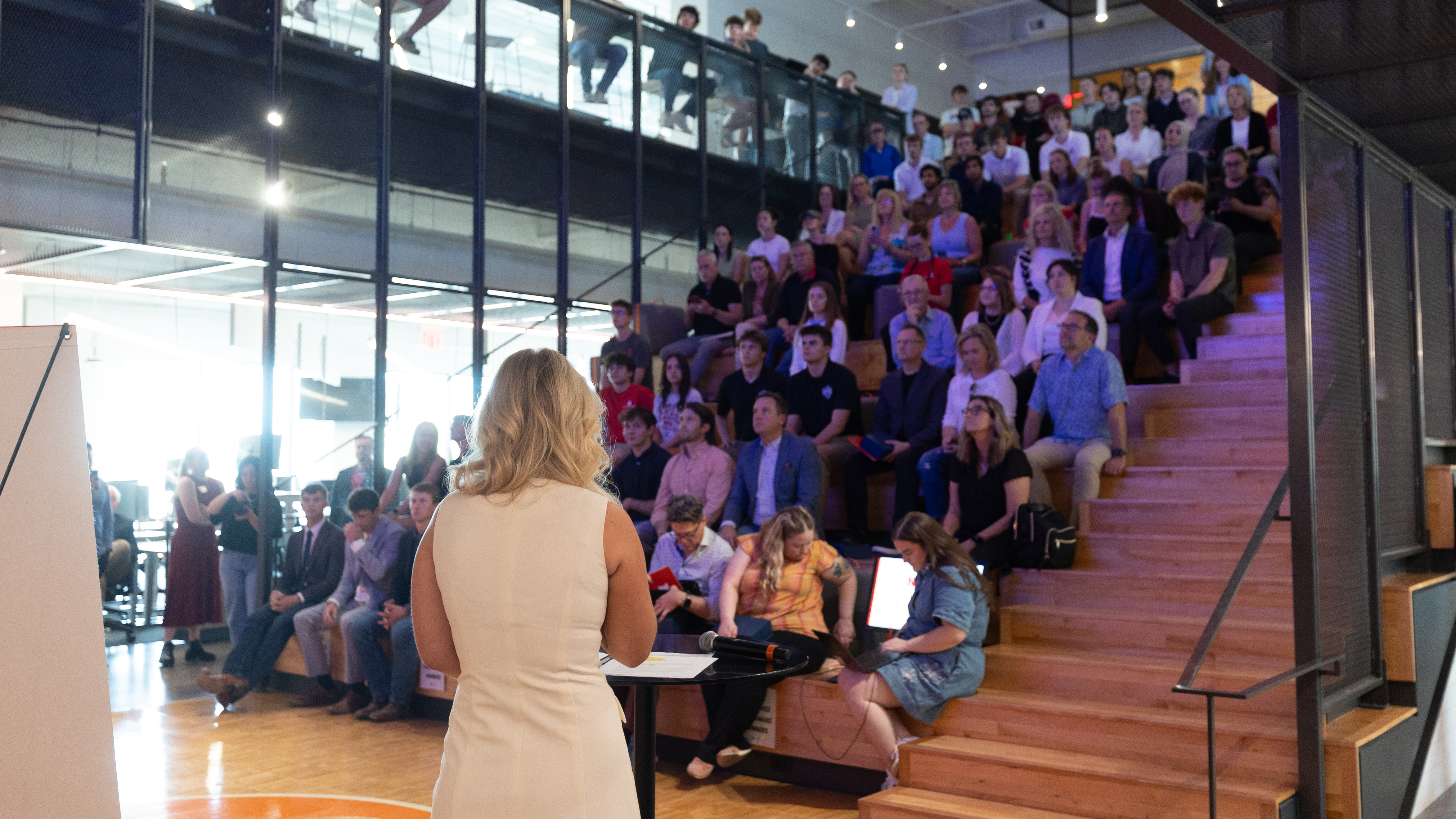 A woman speaks to a crowd seated on bleachers.