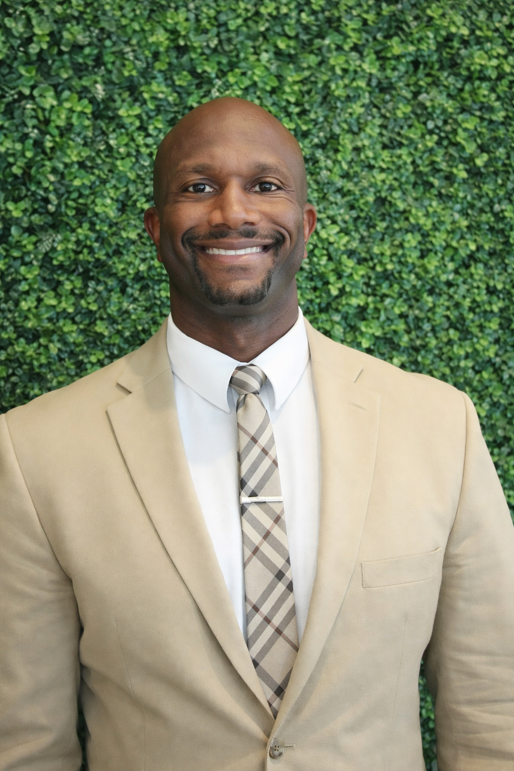 Color portrait of DeMoine Adams, motivational speaker and former Husker football student-athlete. He is wearing a tan sport coat over a white dress shirt with tan tie, in front of a plant wall.