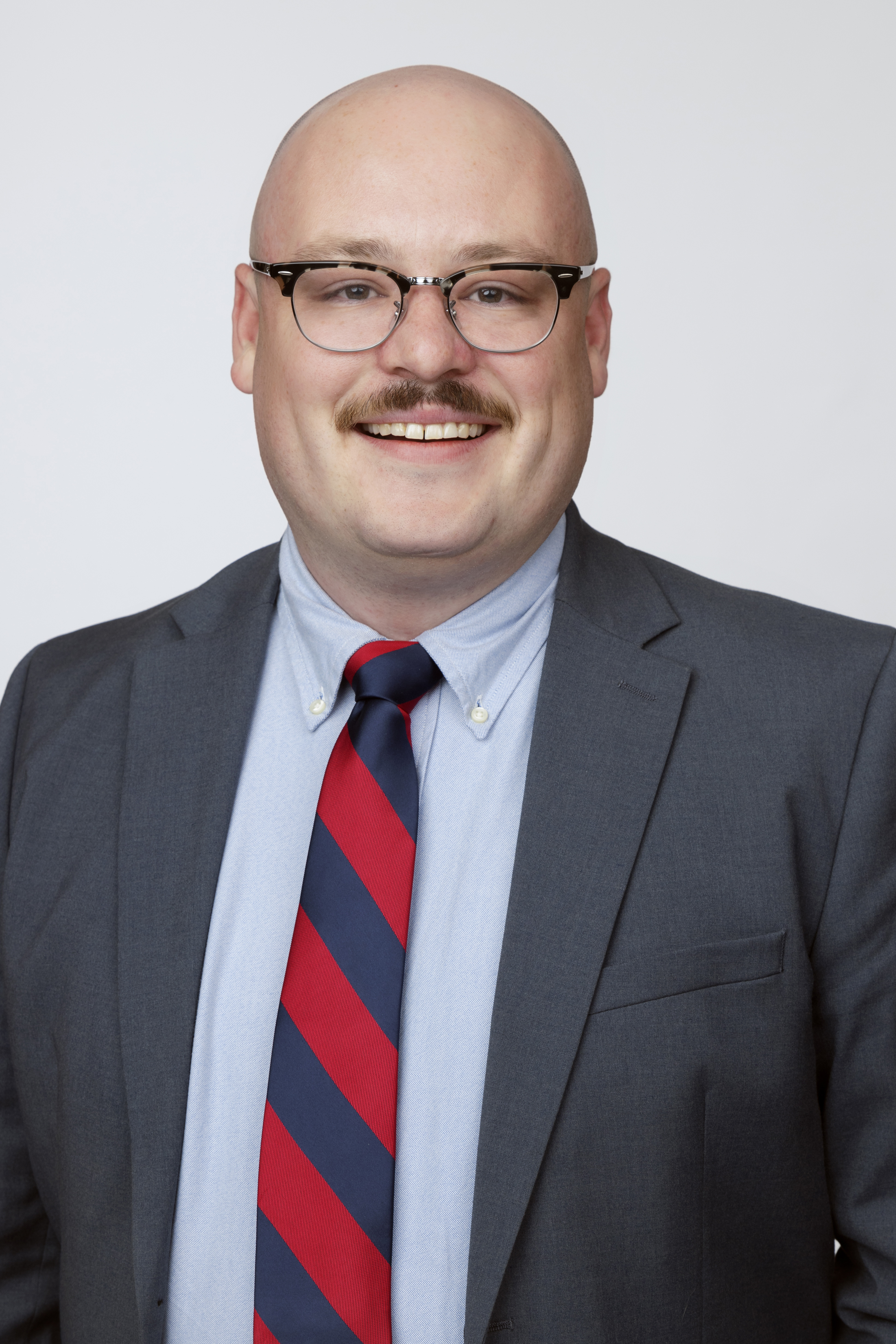 Color portrait of Julian Gillilan, doctoral candidate in the University of Nebraska–Lincoln's Department of History. He is wearing a dark gray sport coat over a light blue dress shirt and red-and-blue striped tie, with glasses.