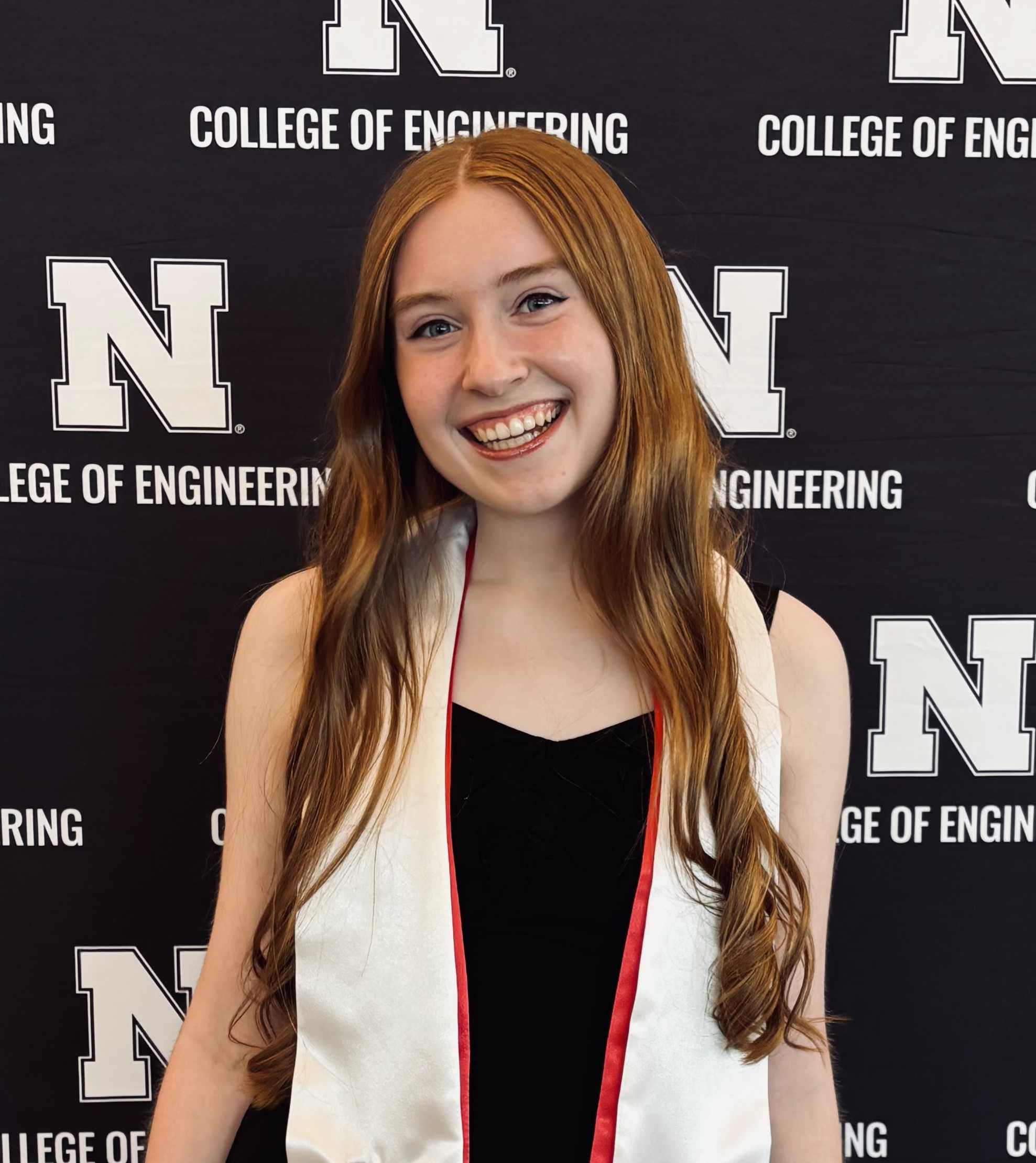 Color portrait of NSF Graduate Research Fellow Gracie Kerr, wearing red and white stoles over a black top, in front of a black-and-white College of Engineering backdrop.