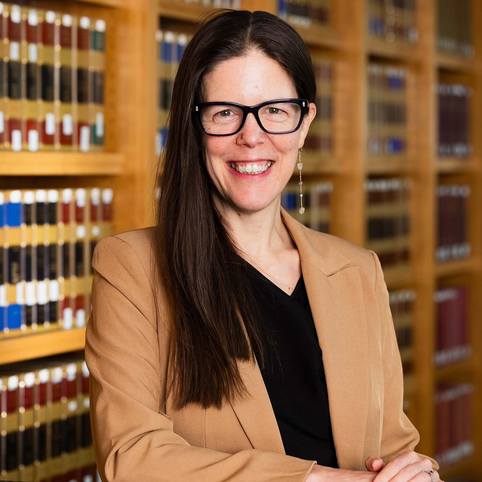 Color portrait of Jessica A. Shoemaker, Steinhart Foundation Distinguished Professor of Law at the University of Nebraska–Lincoln, standing in front of bookcases full of legal tomes. She is wearing a tan blazer over a black V-neck, black-framed glasses and dangly earrings.