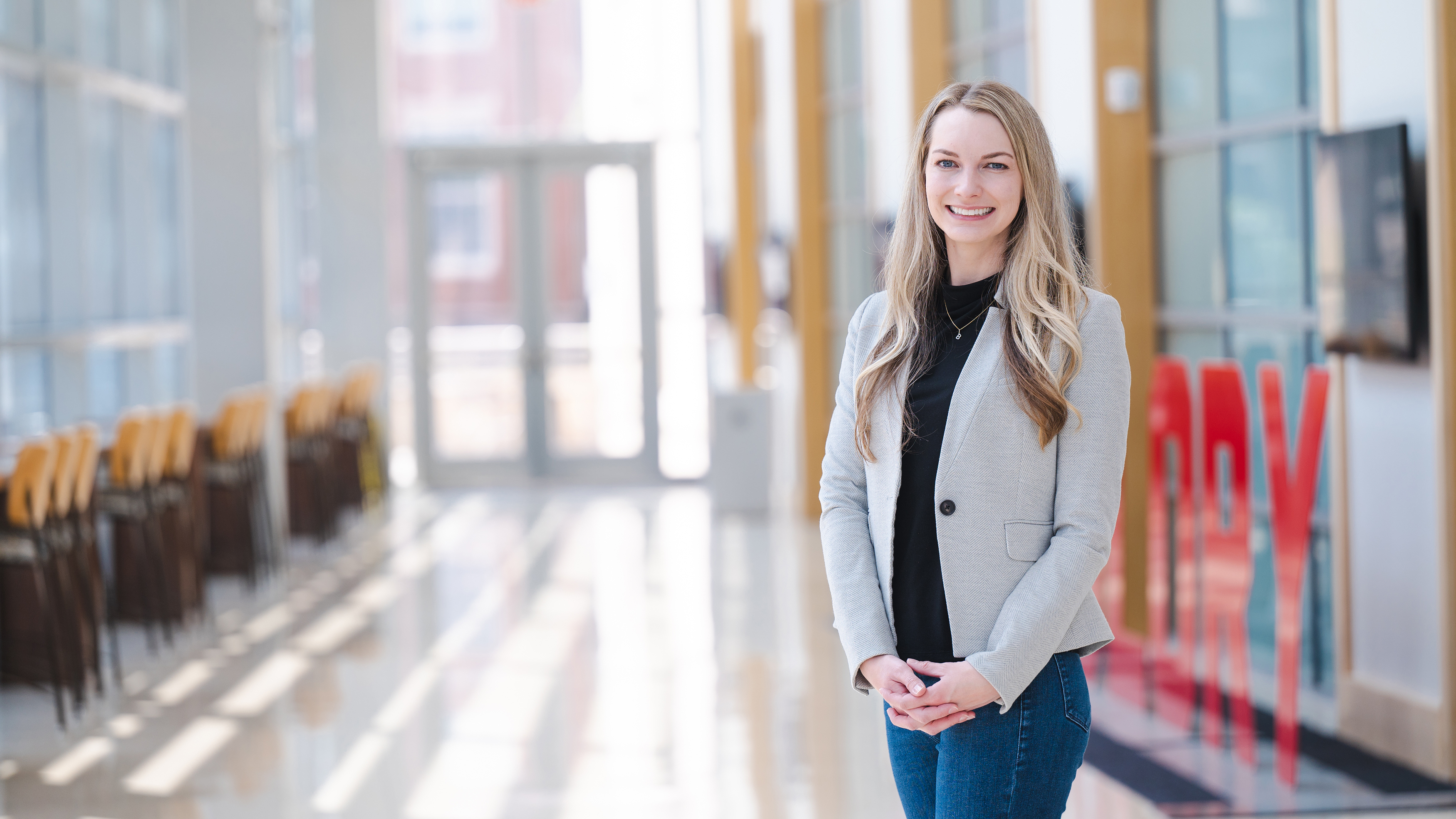 Natalie Chisam, a white blonde woman, stands in a black shirt, grey blazer and blue jeans in the College of Business.