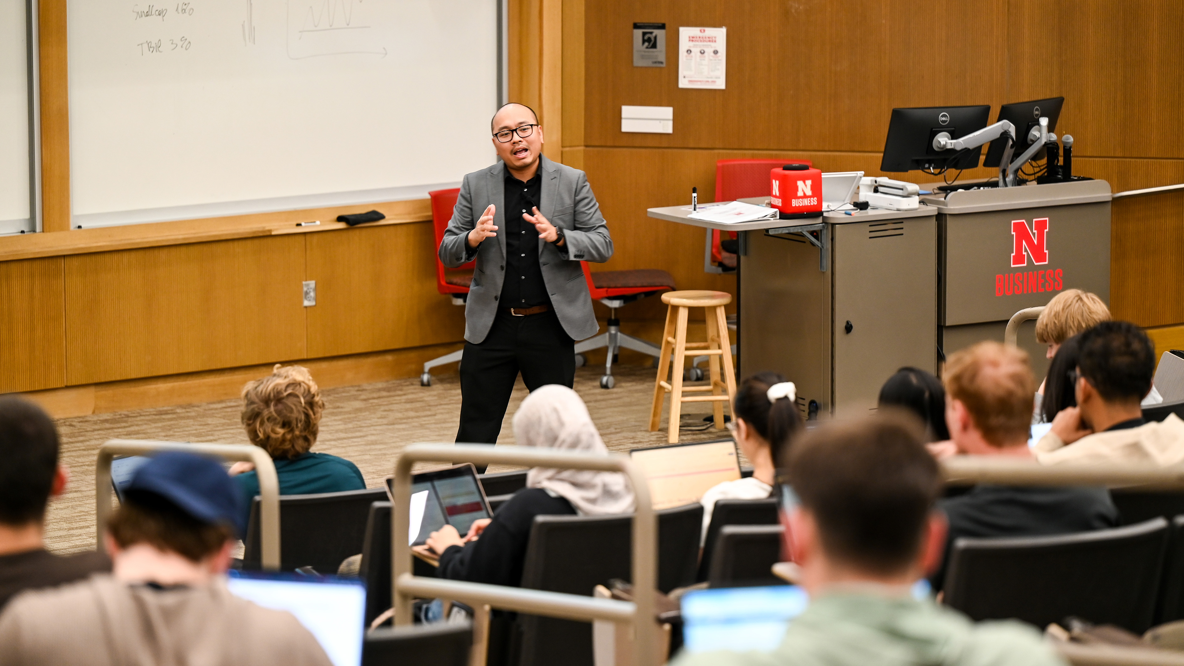A professor wearing a black shirt, slacks and glasses, and a grey blazer lectures a crowd of finance students in front of a white board.