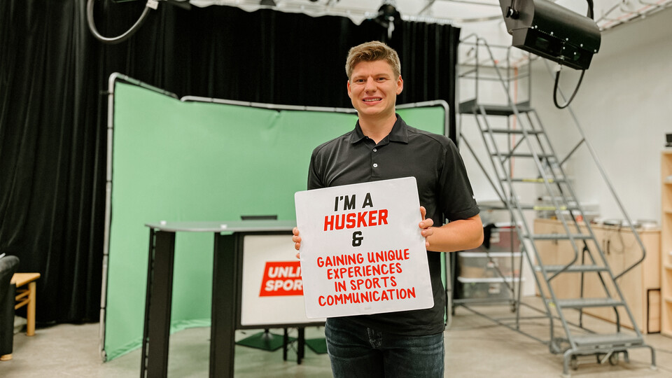 Peyton Thomas stands in front of a green screen and news set holding a sign that reads, "I'm a Husker and gaining unique experiences in sports communication."