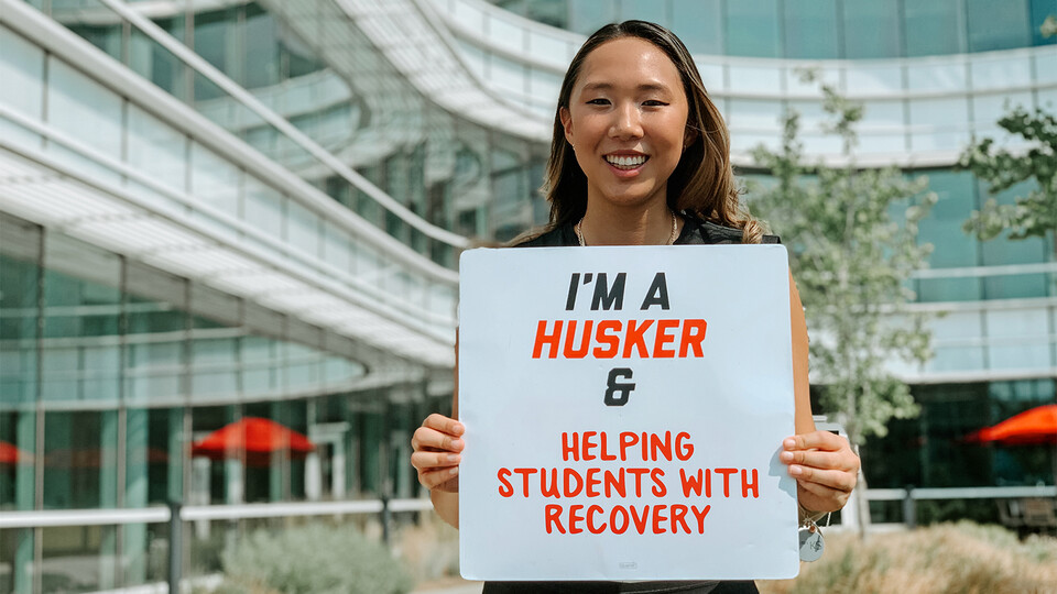 Grace holds up a sign that reads "I'm a Husker & Helping Students with Recovery".