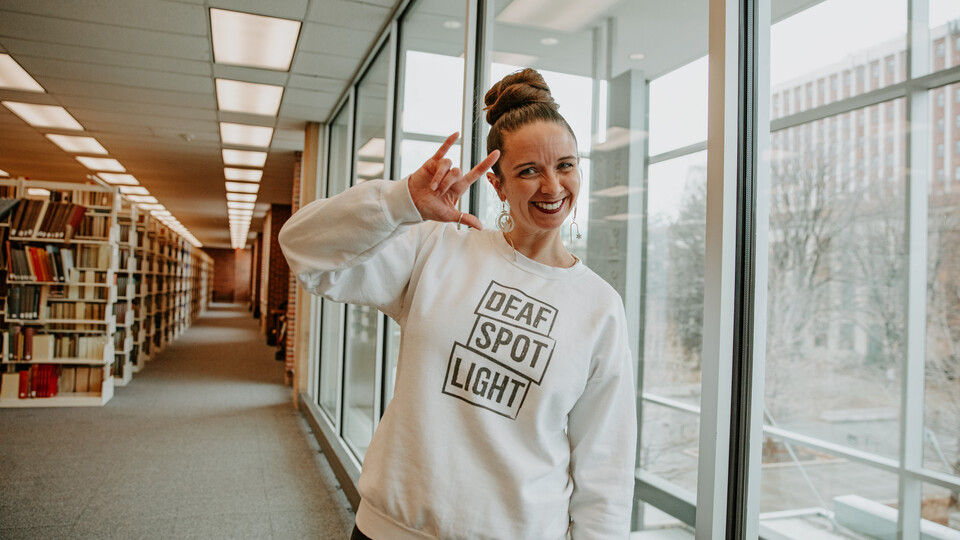 3rd-year PhD candidate Renca Dunn poses in Love Library, wearing a crew neck hoodie that reads "Deaf Spotlight" and signing "I love you."