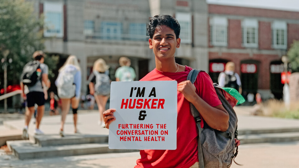Naren smiles for a photo holding a sign that says "I'm a Husker & furthering the conversation on mental health"