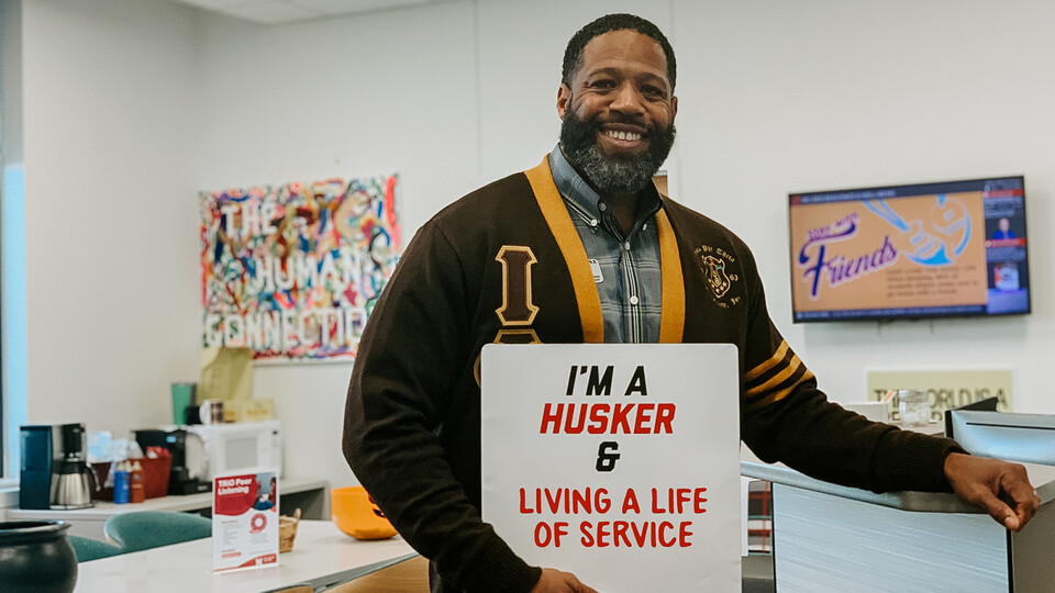 Kenji Madison stands in the Big Red Resilience & Well-Being office holding a sign that reads, "I'm a Husker & Living a Life of Service." 