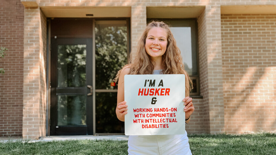 Conradi stands in front of the Barkley Speech-Language & Hearing Clinic on East Campus, holding a sign that reads "I'm a Husker & Working Hands-On with Communities with Intellectual Disabilities”.