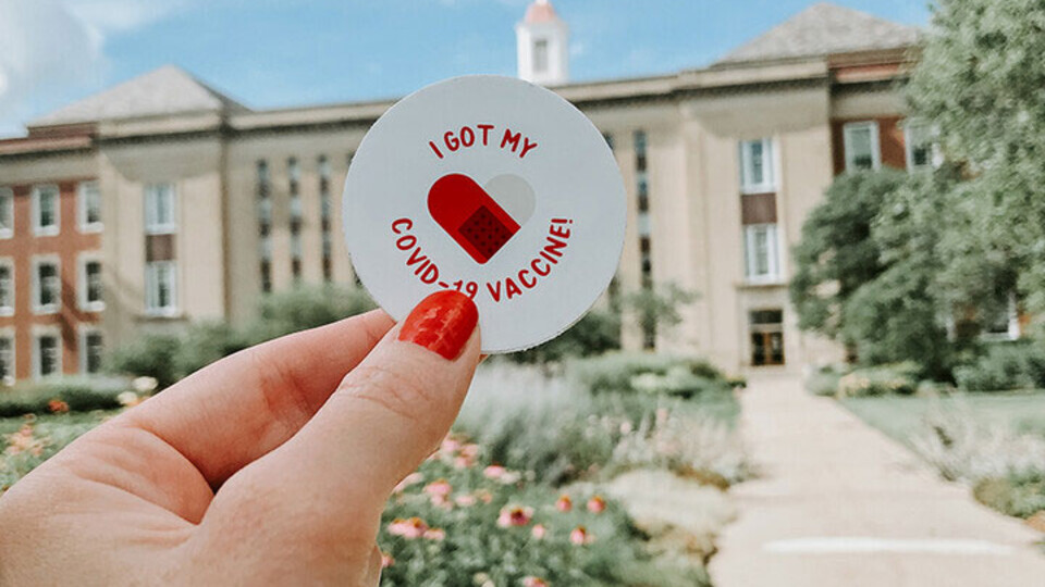 A hand holds an "I've been vaccinated" sticker in the air with the Nebraska Union in the background.