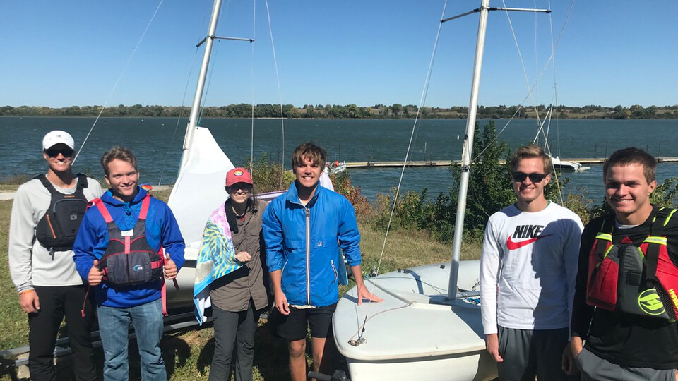 Members of the UNL Sailing Club stand next to their boats while backgrounded by a whitecap-topped lake