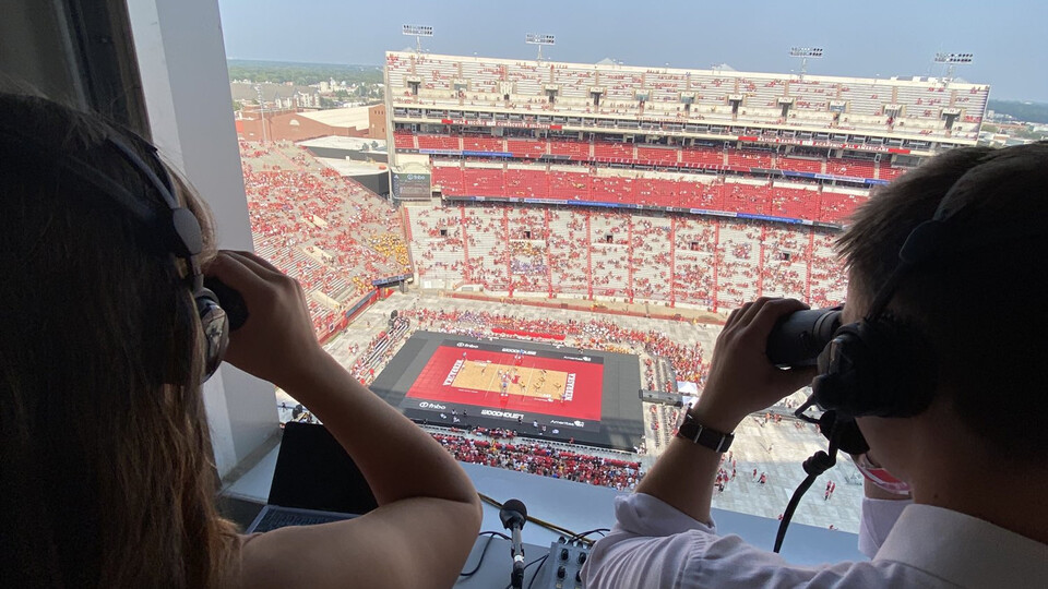 Two students in the press box of Memorial Stadium peer through binoculars at a volleyball match below
