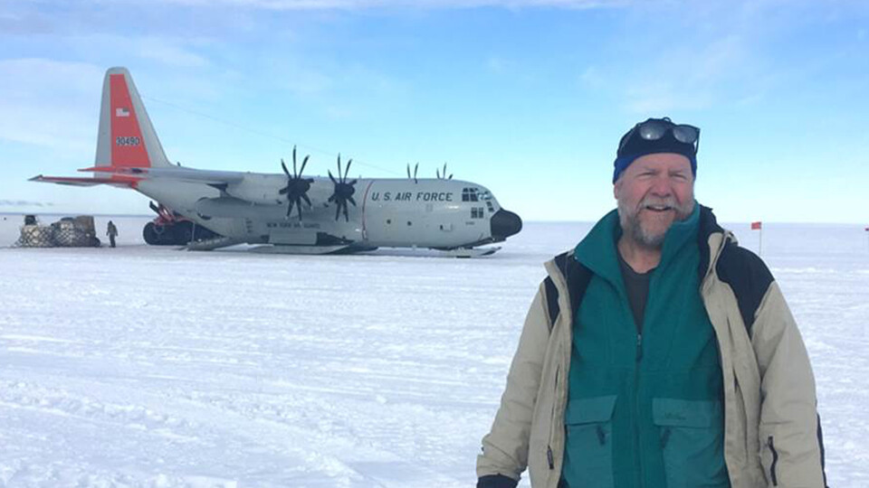 David Harwood stands in the snow of Antarctica with a cargo plane in the background