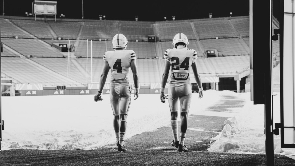 A black-and-white photo of Kamdyn Koch and Nico Ottomanelli, clad in jerseys and helmets, walking onto a snow-covered Tom Osborne Field