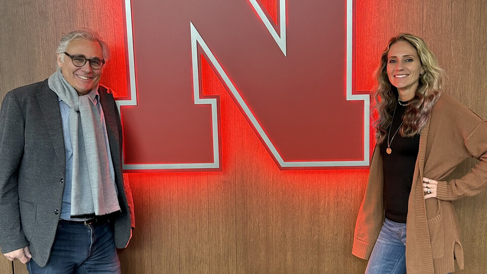 Two visiting professors stand and smile on either side of a large Nebraska N affixed to the wall