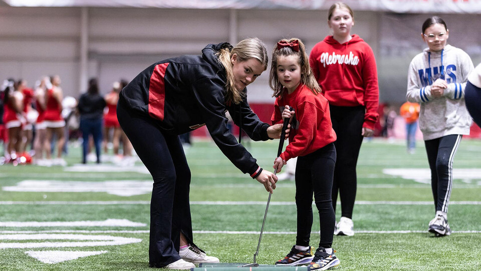 A Husker student-athlete helps a girl putt a golf ball inside the Hawks Championship Center