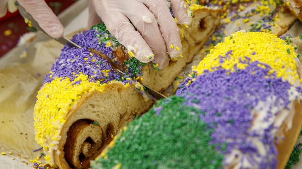 A member of the Dining Services team slices a Kings Cake adorned with yellow, purple and green sprinkles.