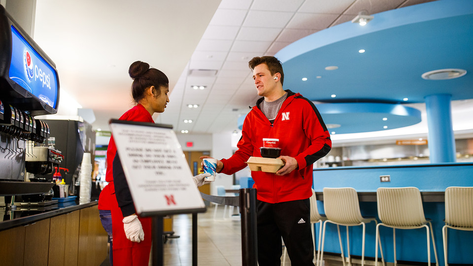 Zachery Borer, a freshman from North Bend, collects a drink and lunch to-go from Manpreet Kaur in the Willa S. Cather Dining Complex on March 17. Cather is one of four dining options that will remain open as of April 4.
