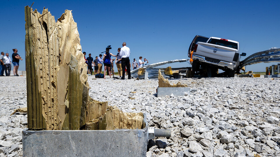A dozen or so people stand next to a pickup that crashed into a metal guardrail, with a splintered wooden post in the foreground, at the Midwest Roadside Safety Facility's Outdoor Proving Grounds.