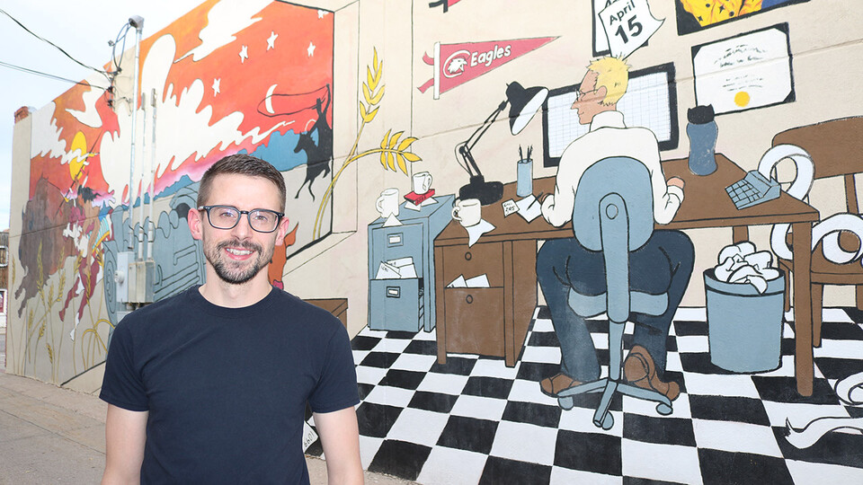 Travis Hencey stands in front of “Beating the Deadline,” a mural he designed for Chadron’s Art Alley. The mural features a man at a desk working on a computer.