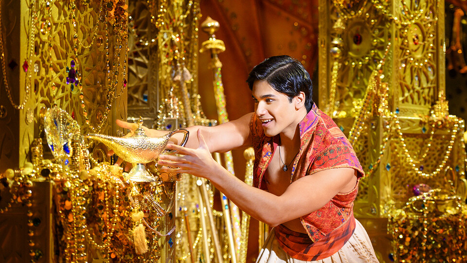 Adi Roy as Aladdin holds a golden lamp in a room full of treasure.