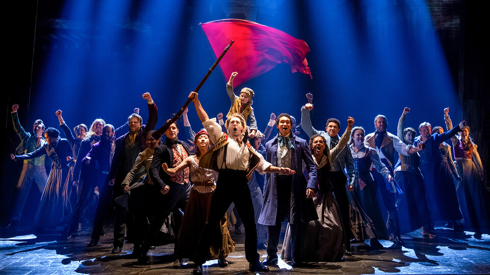 The "Les Miserables" ensemble appears on stage, with a man holding a gun in front and a red flag in the background.