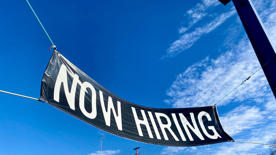 Black "now hiring" sign contrasted against blue sky