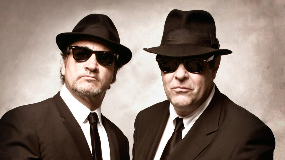 Jim Belushi and Dan Aykroyd, wearing suits with sunglasses and black fedoras