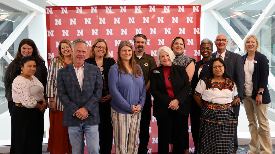 Thirteen people pose for a photograph in two rows, in front of a Husker "N" backdrop.
