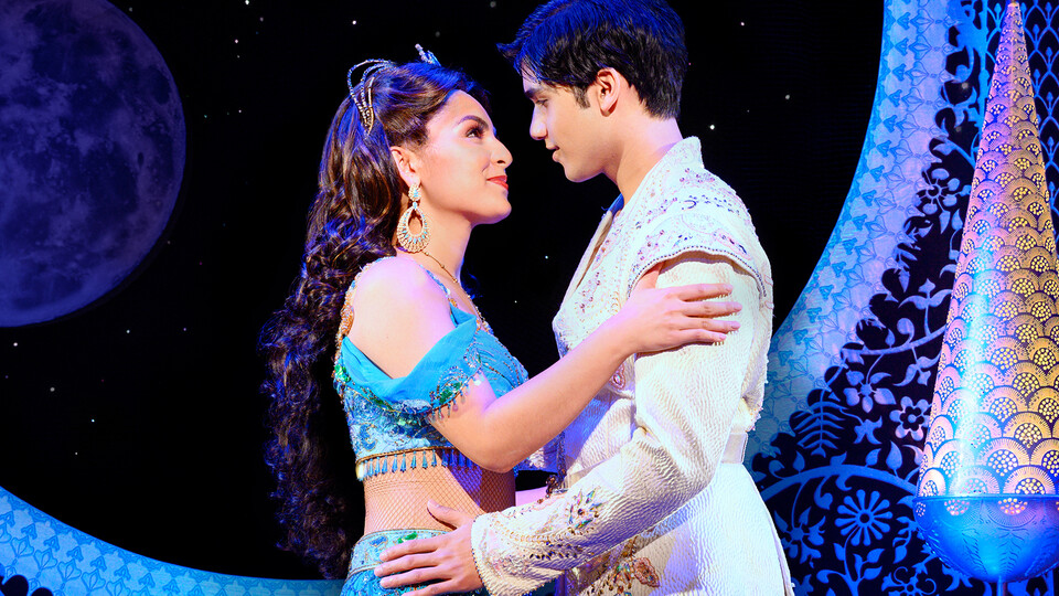 A young woman and young man hold each other and look into each other's eyes in a palace room in "Aladdin."