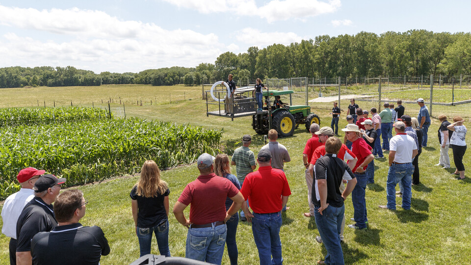 About two dozen people stand near a hail simulator and a corn plot.