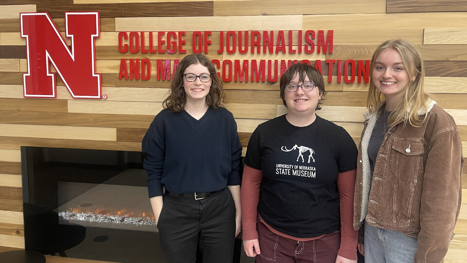 Husker journalists Shelby Rickert, Samantha Grove and Jordan Moore stand in front of an electric fireplace and lettering that read "College of Journalism and Mass Communications."
