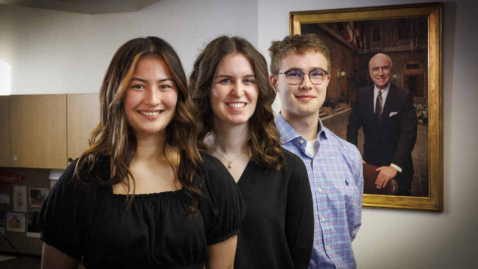Husker undergraduates Kyoko Wall, Victoria Diersen and Zane Mrozla-Mindrup stand in front of a framed portrait of Clayton Yeutter.