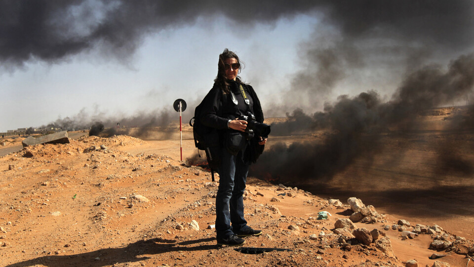 Lynsey Addario stands with a camera on rocky terrain, with plumes of black smoke in the background.