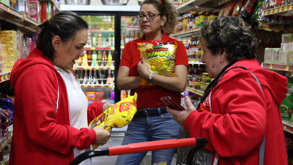 Three women stand around a grocery cart in a store, two holding food packages.