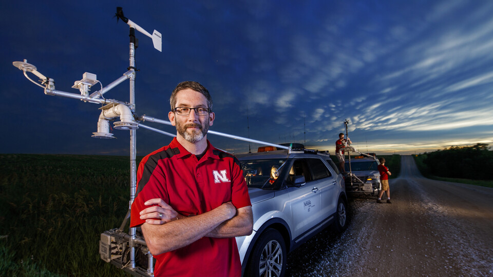 Adam Houston stands with his arms crossed in front of two storm-chasing vehicles.